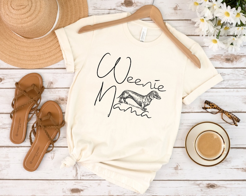 May include: A white t-shirt with the text "Weenie Mama" and a black and white illustration of a dachshund.