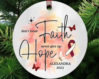 Può includere: Addobbo natalizio rotondo bianco con uno sfondo ad acquerello in tonalità rosa e gialla. L'addobbo presenta le parole "Faith Hope" con il testo "never give up" e "don't loose" in nero. L'addobbo presenta anche un nastro bordeaux, una farfalla e il nome "ALEXANDRA 2023".