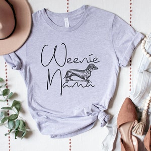 May include: A light gray t-shirt with the text "Weenie Mama" in black script. A black and white illustration of a dachshund is centered below the text.
