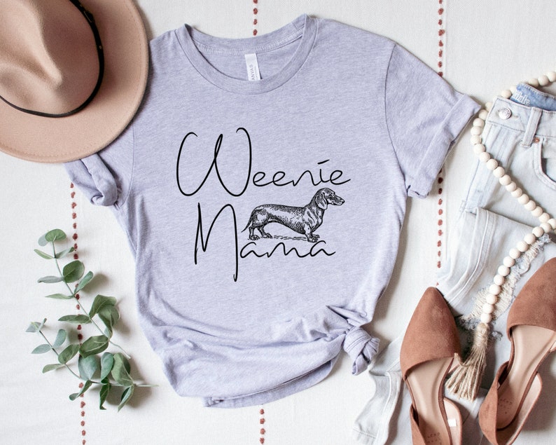 May include: A light gray t-shirt with the text "Weenie Mama" in black script. A black and white illustration of a dachshund is centered below the text.