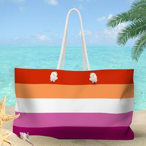 Peut inclure: Un sac fourre-tout rayé rouge, orange, blanc, rose et bordeaux avec des anses en corde blanche. Le sac est sur une plage de sable avec une mer bleue et un palmier en arrière-plan.
