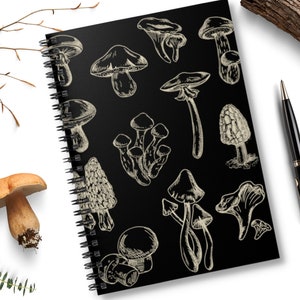 May include: A black spiral notebook with a white background featuring a variety of mushroom illustrations. The mushrooms are drawn in a vintage style and are arranged in a random pattern.