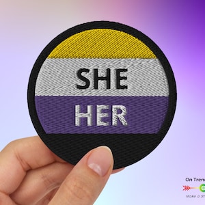 May include: A round embroidered patch with the words "SHE HER" in white text on a purple, white, and yellow background. The patch is surrounded by a black border.