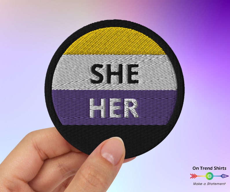以下が含まれることがあります： 紫、白、黄色の背景に白い文字で「SHE HER」と書かれた丸い刺繍パッチ。パッチは黒い縁取りで囲まれている。