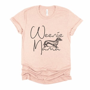May include: A light pink t-shirt with the text "Weenie Mama" in black script. A black and white illustration of a dachshund is between the words.