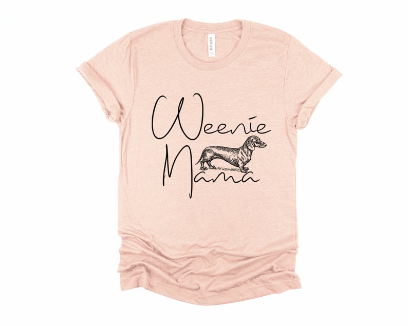 May include: A light pink t-shirt with the text "Weenie Mama" in black script. A black and white illustration of a dachshund is between the words.