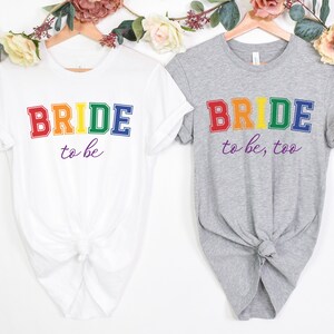 Puede incluir: Dos camisetas blancas con el texto "BRIDE to be" y "BRIDE to be, too" en colores arcoíris. El texto está en negrita, con una fuente de bloque.