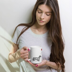 Puede incluir: Taza de cer&aacute;mica blanca con una ilustraci&oacute;n en blanco y negro de un dachshund sosteniendo un globo rojo. La taza est&aacute; siendo sostenida por una persona.
