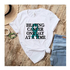 May include: White t-shirt with a teal ribbon and the text "BEATING CANCER ONE DAY AT A TIME" in black.