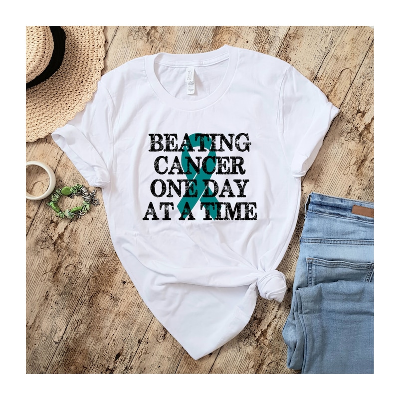 May include: White t-shirt with a teal ribbon and the text "BEATING CANCER ONE DAY AT A TIME" in black.