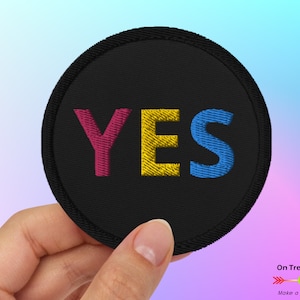 Peut inclure: Un patch rond noir brodé avec le mot "YES" en fil rose, jaune et bleu.