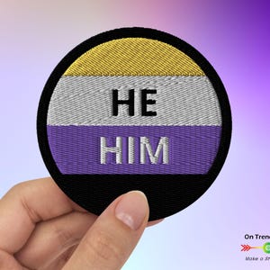 May include: A round embroidered patch with the non-binary pride flag colors. The patch has a black border and the words "HE HIM" in white text.