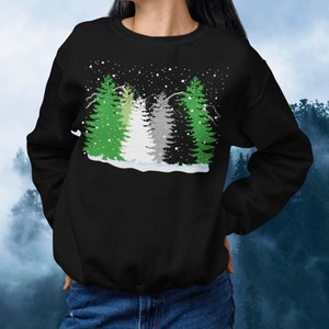 May include: Black sweatshirt with a winter scene of green, white, and gray trees with snowflakes falling. The trees are stylized to resemble a pride flag.