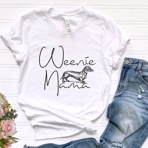 May include: White t-shirt with the text "Weenie Mama" in black script font. A black and white illustration of a dachshund is centered below the text.