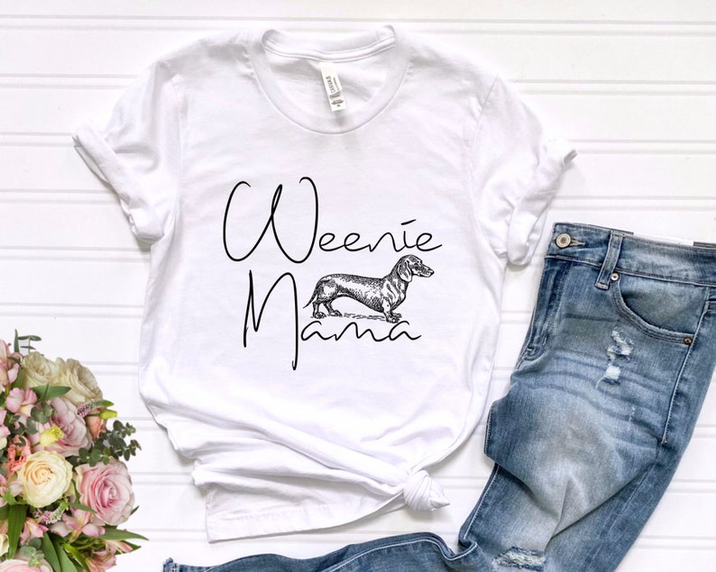 May include: White t-shirt with the text "Weenie Mama" in black script font. A black and white illustration of a dachshund is centered below the text.