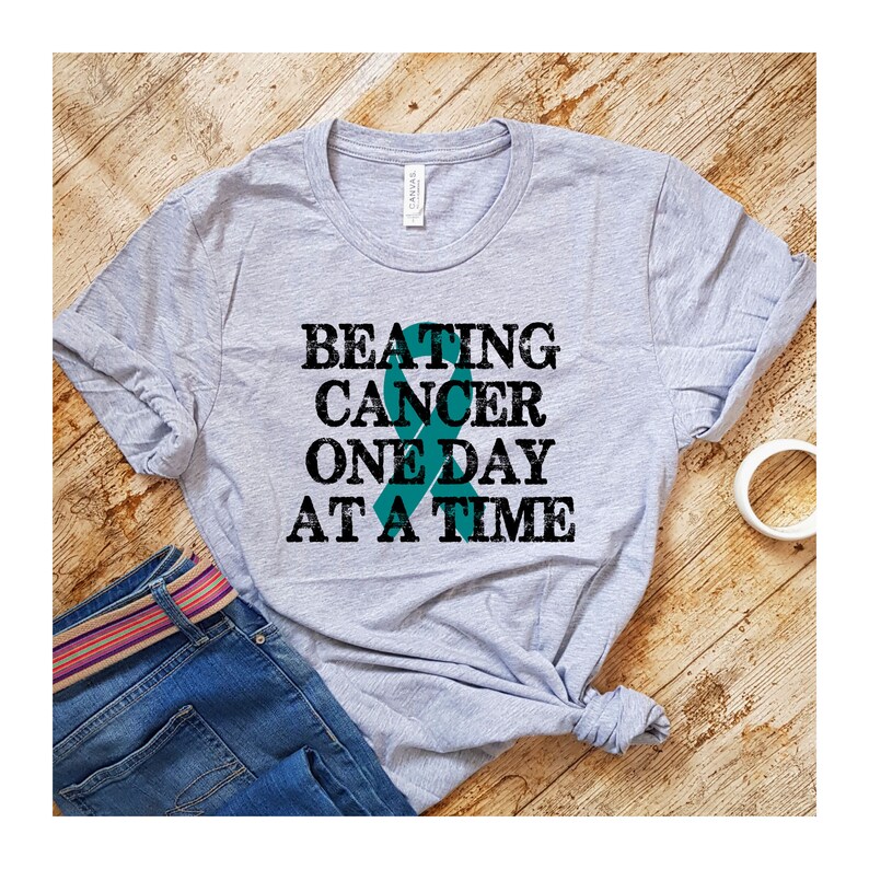 May include: A grey t-shirt with the text "BEATING CANCER ONE DAY AT A TIME" in black. A teal ribbon is behind the text.