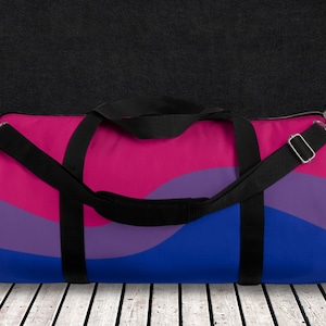 May include: A black duffel bag with a pink, purple and blue wavy pattern. The bag has a black shoulder strap and two black handles.