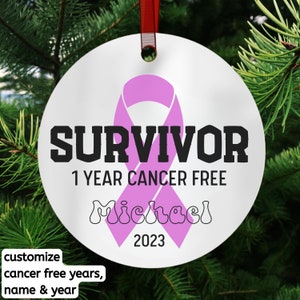May include: A white round Christmas ornament with a pink ribbon and the text "SURVIVOR 1 YEAR CANCER FREE Michael 2023".