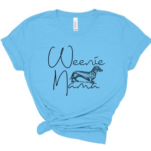 May include: A light blue t-shirt with the text "Weenie Mama" in black script. A black and white illustration of a dachshund is centered below the text.