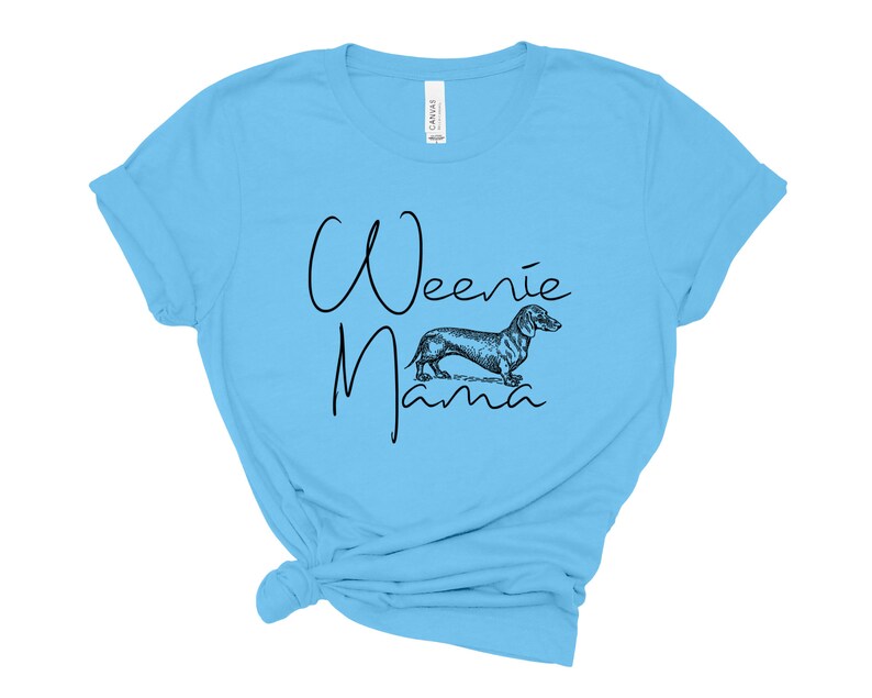 May include: A light blue t-shirt with the text "Weenie Mama" in black script. A black and white illustration of a dachshund is centered below the text.