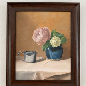 Puede incluir: Pintura de naturaleza muerta enmarcada que presenta un jarrón azul con flores rosas y blancas, una taza plateada y un mantel blanco. El fondo es de color marrón dorado cálido, y el marco es marrón oscuro.