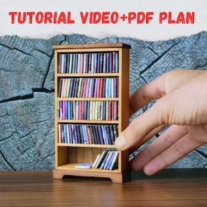 Peut inclure: Une petite bibliothèque en bois remplie de livres miniatures colorés. La bibliothèque est tenue par une main. Le texte "TUTORIAL VIDEO+PDF PLAN" est en haut. La bibliothèque est en bois brun clair et comporte trois étagères.