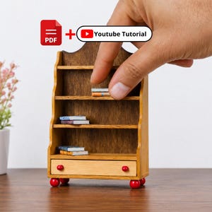 May include: A miniature wooden bookshelf with three open shelves and a bottom drawer featuring two small red knobs. The unit stands on four red spherical feet. Several tiny books are placed on the shelves. Includes PDF and Youtube Tutorial text.