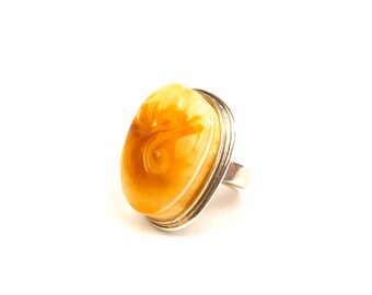 May include: Oval-shaped amber ring with a silver band. The amber stone has a light brown color with a swirl design. The ring is set on a white background.