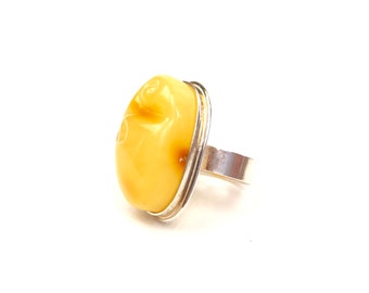 May include: A silver ring featuring a large, oval-shaped amber stone. The amber has a creamy yellow color with subtle swirls and carved details. The ring's band is a simple, polished silver.