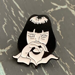 May include: Black and white enamel pin featuring a stylized illustration of a woman with black hair and a white shirt, covering her mouth with her hand.