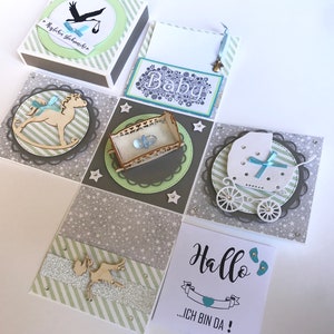 Può includere: Una scatola esplosiva fatta a mano a tema baby con vari elementi decorativi. La scatola presenta una cicogna, un cavallo a dondolo, un passeggino e una culla. La combinazione di colori include grigio, bianco e verde chiaro, con accenti di blu. La scatola ha anche la parola "Baby" e il testo "Hallo...ICH BIN DA!"