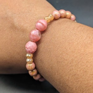Puede incluir: Un brazalete de cuentas rosa y dorado con cuentas facetadas. El brazalete está hecho con una mezcla de cuentas redondas y ovaladas.