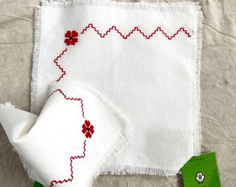 May include: Two white linen napkins with red embroidered flowers and a scalloped edge. The napkins are fringed and have a white tag with black text.