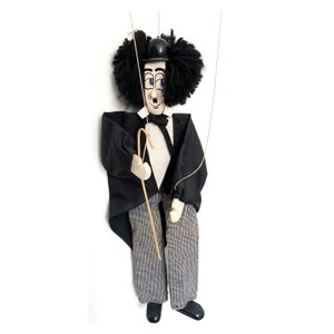 Puede incluir: Un títere de madera de Charlie Chaplin, con un traje negro, una pajarita negra y un bombín negro. Tiene un bigote negro y un bastón en la mano.