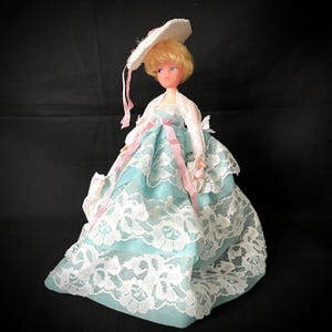 Puede incluir: Muñeca vintage con un vestido azul claro con encaje blanco superpuesto. La muñeca tiene cabello rubio, ojos azules y un sombrero blanco con cintas rosas. La muñeca lleva una chaqueta bolero blanca.