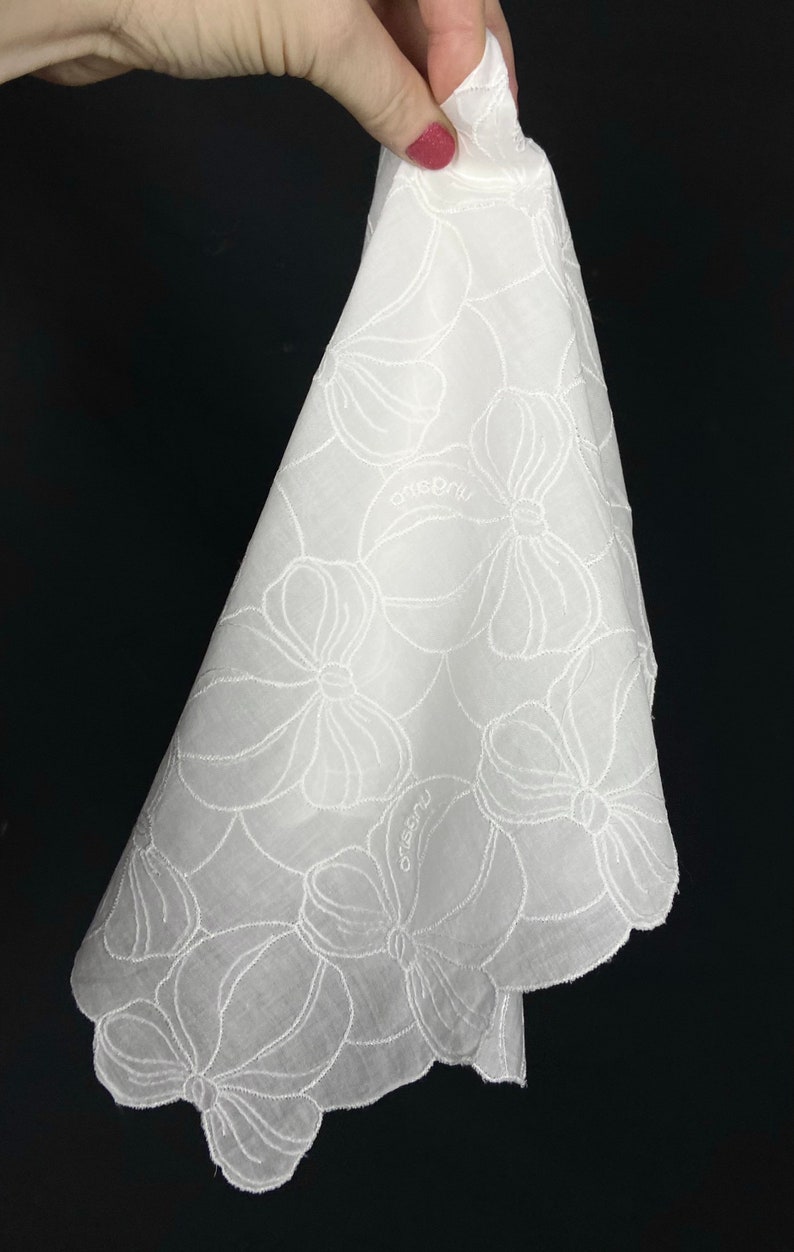 May include: White cotton handkerchief with a scalloped edge and embroidered floral design. The handkerchief has the word "ocrebru" embroidered on it.