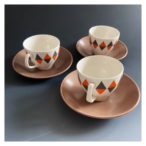 May include: A set of three vintage teacups and saucers. The cups are white with a geometric pattern of orange and grey diamonds. The saucers are a solid, muted brown colour. The cups have a curved handle.