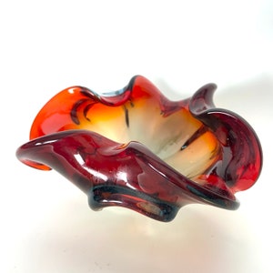 May include: A red, orange and white glass bowl with a wavy, organic shape. The bowl is made of clear glass with a red, orange and white swirl pattern.