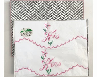 May include: A white pillowcase set with pink embroidery. The pillowcases feature pink flowers and the words "His" and "Hers" embroidered on them. The pillowcases are packaged in a white box with a black and white chevron pattern.