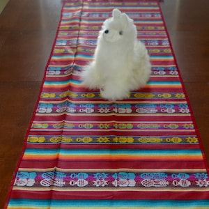 May include: A white llama plush toy sitting on a colorful striped table runner with a fringe border. The runner has a repeating pattern of pineapples and fish.