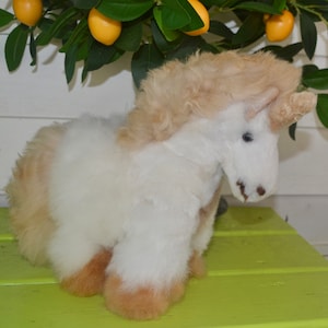 May include: A white and tan fluffy unicorn stuffed animal with a brown horn. The unicorn is sitting on a green surface.