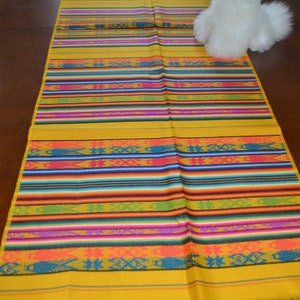 May include: A yellow table runner with colorful stripes and a woven pattern. The runner has fringe on the ends.