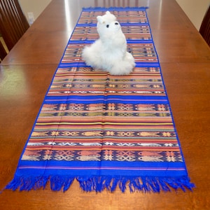 May include: A blue and multi-colored table runner with fringe on the ends. The runner has a woven pattern of fish and geometric designs. A white stuffed animal is sitting on the runner.