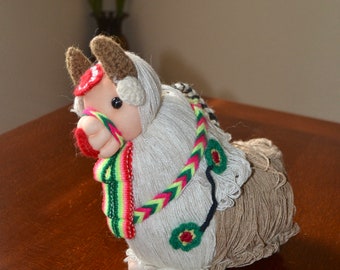 May include: A white and brown crocheted llama toy with a red, green, and yellow striped scarf and a brown head. The llama is sitting on a wooden surface.