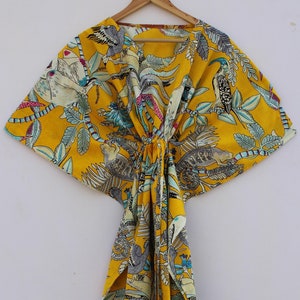May include: A yellow, patterned kaftan dress with a tropical bird and foliage design. The dress is made of lightweight fabric and hangs from a wooden hanger. The design includes birds, leaves, and branches in shades of gray, blue, and pink.