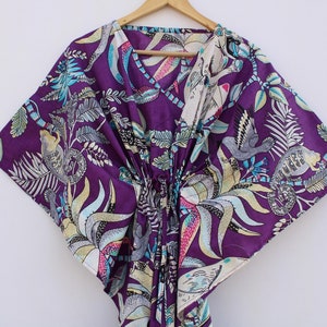 May include: A purple kaftan with a V-neck and a drawstring waist. The fabric features a colorful print of birds, monkeys, and foliage in shades of blue, green, and cream. The garment is hanging on a wooden hanger.