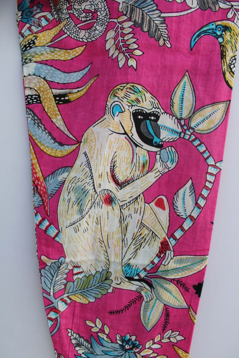 Peut inclure: Tissu rose vif avec une illustration de singe blanc, un visage bleu et un museau noir, tenant un fruit. Le motif comprend des feuilles color&eacute;es et un oiseau sur fond rose.