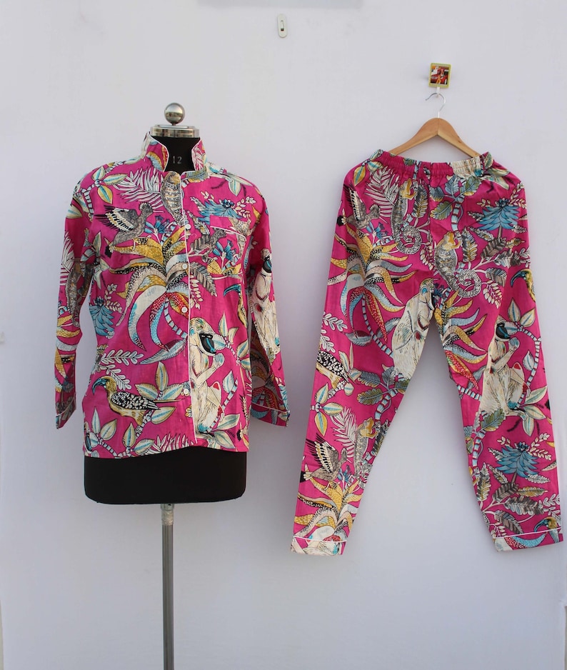 Peut inclure: Un ensemble de pyjama rose vif, orn&eacute; d'un imprim&eacute; color&eacute; de motifs botaniques et d'oiseaux. L'ensemble comprend une chemise boutonn&eacute;e &agrave; manches longues et un pantalon assorti. La chemise est pr&eacute;sent&eacute;e sur un mannequin, le pantalon sur un cintre en bois.