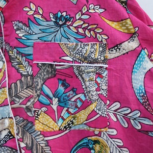 Peut inclure: Gros plan sur un haut de pyjama rose avec un imprim&eacute; floral et d'oiseaux vibrant. Le motif pr&eacute;sente des accents turquoise, jaunes et gris, avec une bordure blanche autour de la poche et du col. Le tissu est l&eacute;ger et doux.