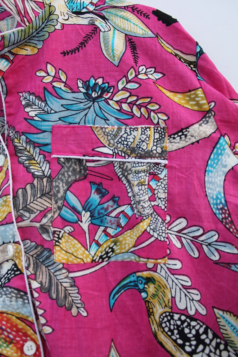 Peut inclure: Gros plan sur un haut de pyjama rose avec un imprim&eacute; floral et d'oiseaux vibrant. Le motif pr&eacute;sente des accents turquoise, jaunes et gris, avec une bordure blanche autour de la poche et du col. Le tissu est l&eacute;ger et doux.